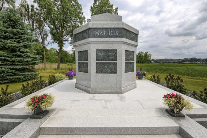Gray granite mausoleum with the name “MATHEIS” engraved at the top, set on a raised stone platform with flower planters and surrounded by trees and open countryside.